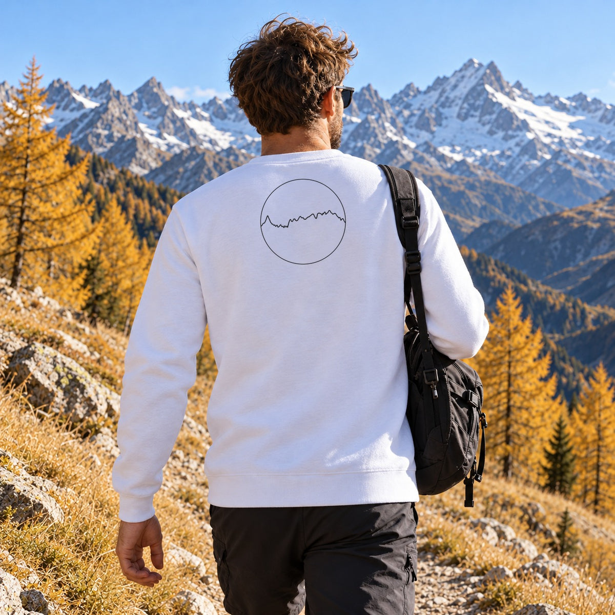 Sweat blanc Les Alpes Kapitales porté de dos sur un sentier d'alpage en automne, médaillon du profil des Alpes dans le dos