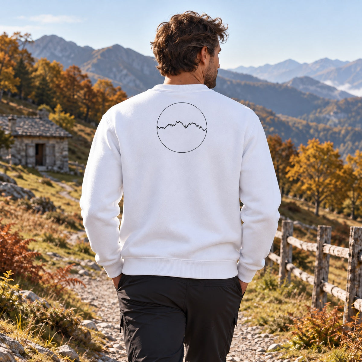 Sweat blanc Les Pyrénées Kapitales porté de dos dans la rue d'un village de montagne en automne, médaillon du profil des Pyrénées dans le dos