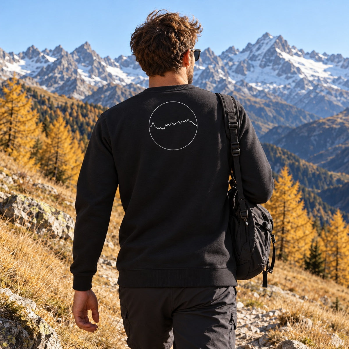 Sweat noir Les Alpes Kapitales porté de dos sur un sentier d'alpage en automne, médaillon du profil des Alpes dans le dos