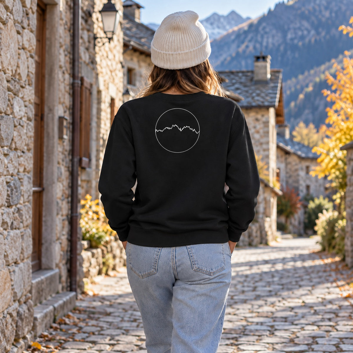 Sweat noir Les Pyrénées Kapitales porté de dos dans un village de montagne en automne, médaillon du profil des Pyrénées dans le dos