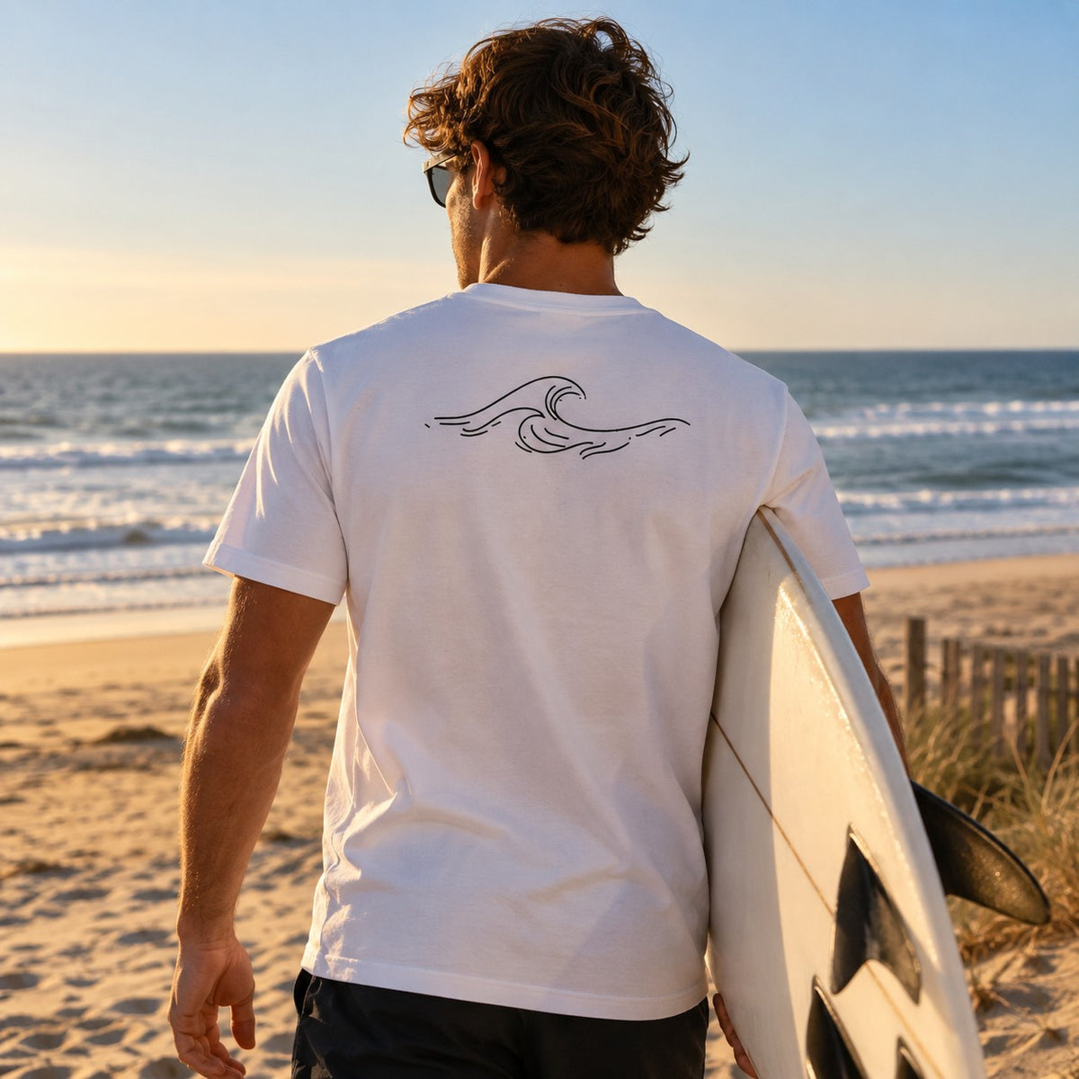 T-shirt blanc Côte Atlantique Kapitales porté de dos face à l'océan, planche de surf sous le bras, vague dessinée dans le dos